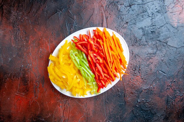 Top view colorful cut peppers on white plate on dark red table free space