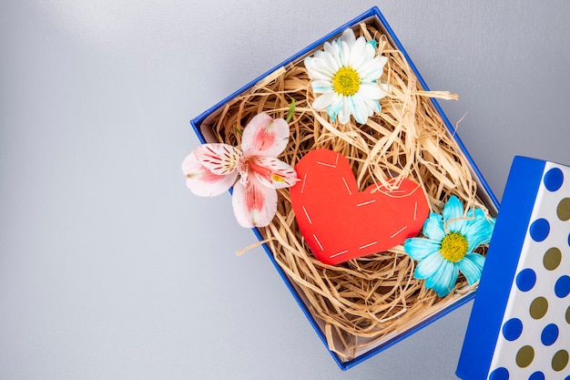 カラフルなデイジーの花とピンクのアルストロメリアの赤い色の紙から作られた心とコピースペースを持つ白いテーブルの青いプレゼントボックスにストローのトップビュー