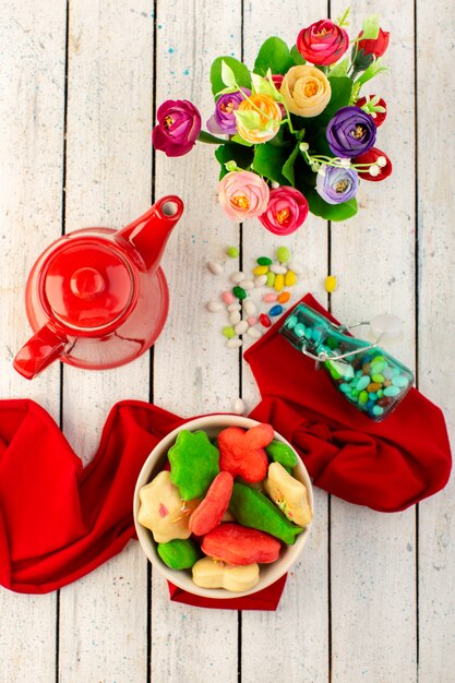 Top view of colorful delicious cookies different formed inside plate with red kettle, candies and flowers