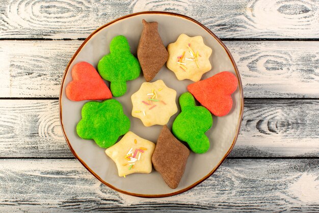 Top view of colorful delicious cookies different formed inside round plate on the grey wooden desk