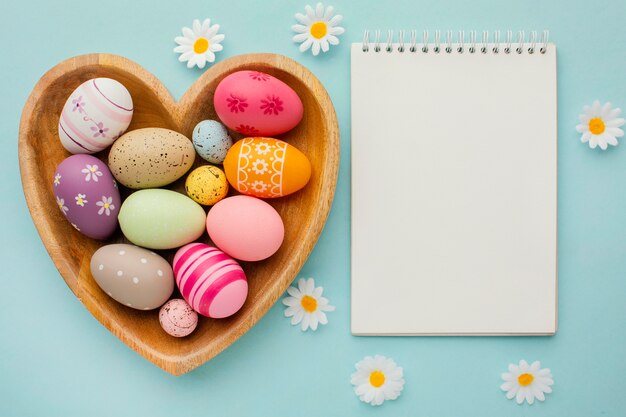 Top view of colorful easter eggs in heart-shaped plate with notebook and flowers