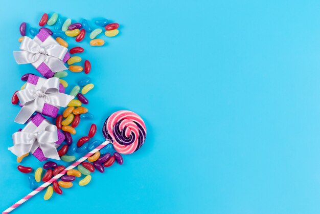 A top view colorful lollipop with little marmalades and purple gift boxes on blue, sugar confectionery candy