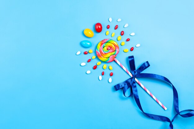 A top view colorful lollipop with pink-white stick blue bow and multicolored candies on the blue background birthday celebration party