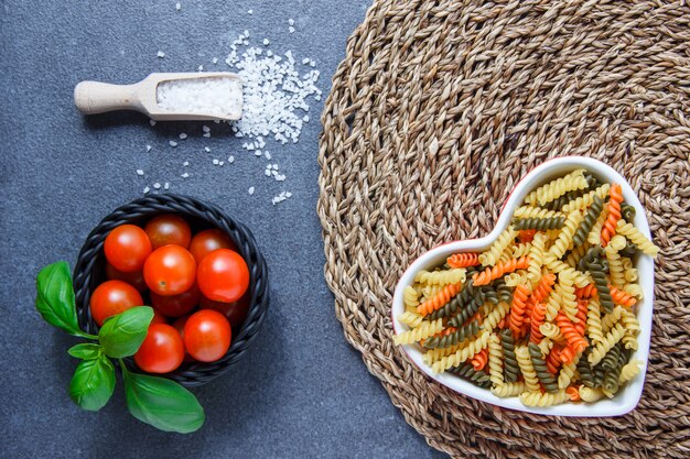 Top view colorful macaroni pasta in heart shaped bowl with tomatoes, leaves, crystal salt on trivet and gray surface. horizontal