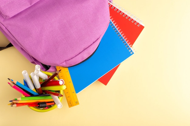 Top view colorful pencils with copybooks and bag on light-yellow wall school felt pen pencil book notepad