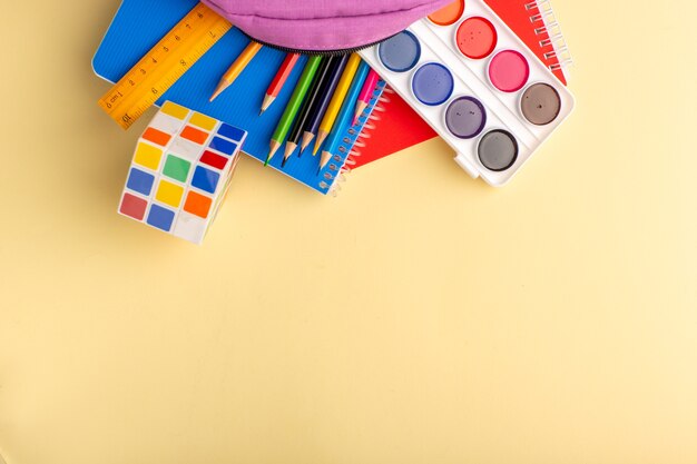 Top view colorful pencils with copybooks paints and purple bag on light-yellow desk school felt pen pencil book notepad