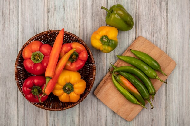 Top view of colorful peppers on a bucket with long shape peppers on a wooden kitchen board on a grey wooden background