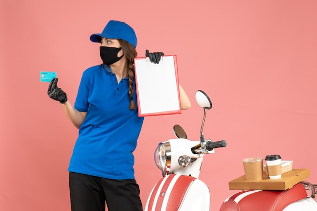 Top view of concentrated courier girl wearing medical mask gloves standing next to motorcycle with coffee cake on it holding documents bank card on pastel peach color background