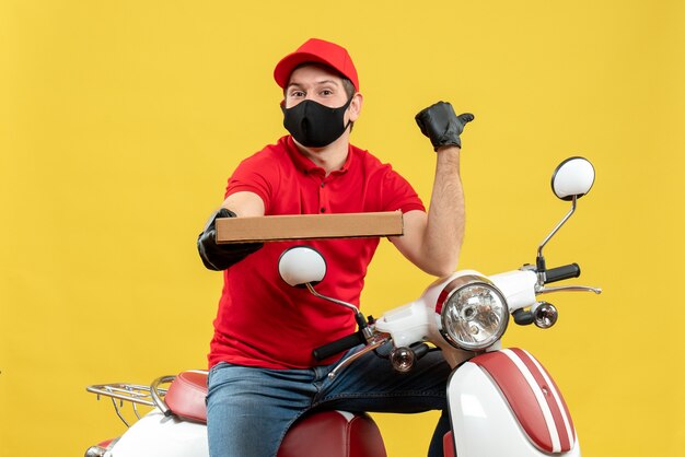 Top view of concentrated courier man wearing red blouse and hat gloves in medical mask sitting on scooter showing order pointing back