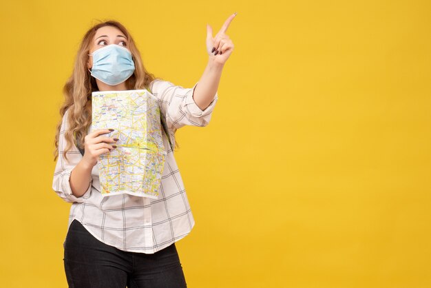 Top view of concentrated travelling girl wearing her mask and backpack holding map pointing up on yellow 