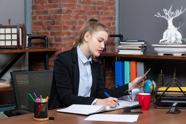 Top view of concentrated young woman sitting at a table and writing on a document in the office