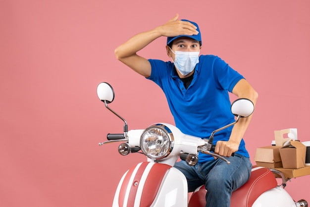 Top view of concerned delivery guy in medical mask wearing hat sitting on scooter on pastel peach background