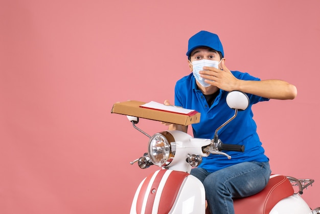 Top view of concerned male delivery person in mask wearing hat sitting on scooter delivering orders holding document on peach background