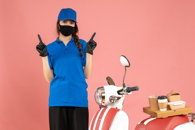 Top view of confident courier girl wearing medical mask gloves standing next to motorcycle with coffee cake on it pointing both sides on pastel peach color background