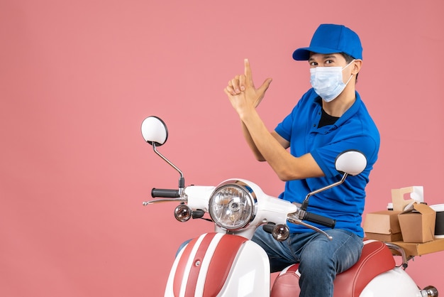 Top view of confident courier man in medical mask wearing hat sitting on scooter making gun gesture on pastel peach background