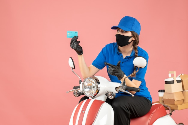 Top view of confident courier woman wearing medical mask and gloves sitting on scooter holding bank card delivering orders on pastel peach background