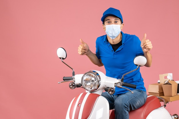 Вид сверху уверенного доставщика в медицинской маске в шляпе, сидящего на скутере и делающего жест на пастельном персиковом фоне