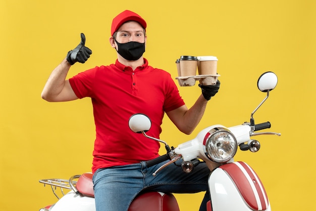 Top view of confident delivery guy wearing uniform and hat gloves in medical mask sitting on scooter showing orders with thumb up