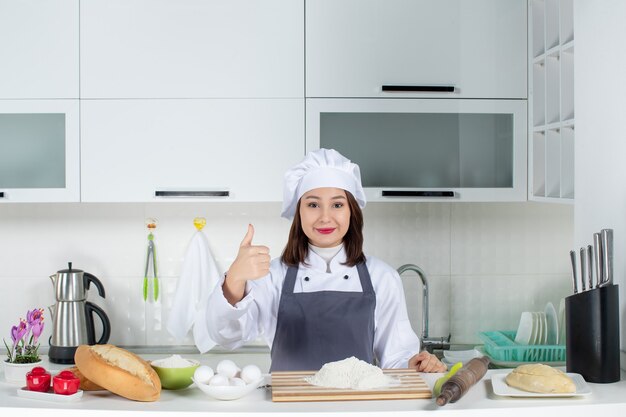 白いキッチンでまな板のパン野菜がOKジェスチャーをしているテーブルの後ろに立っている制服を着た自信のある女性シェフの上面図