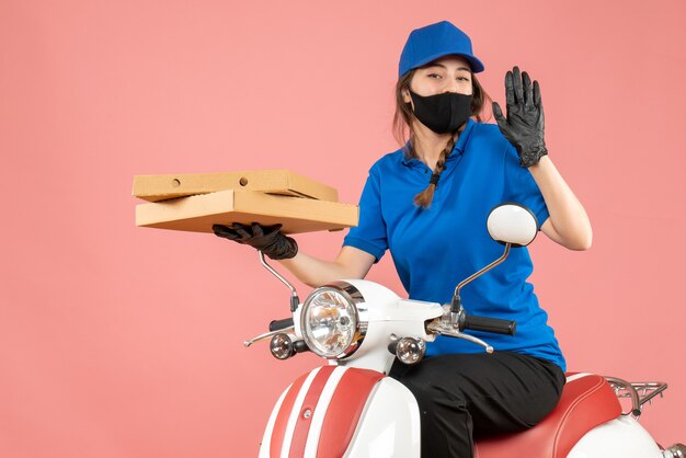 Вид сверху уверенной женщины-курьера в медицинской маске и перчатках, сидящей на скутере, доставляющей заказы на пастельно-персиковом фоне
