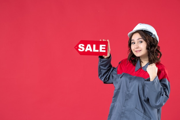 Top view of confident female worker in uniform wearing hard hat and pointing sale icon on isolated red background