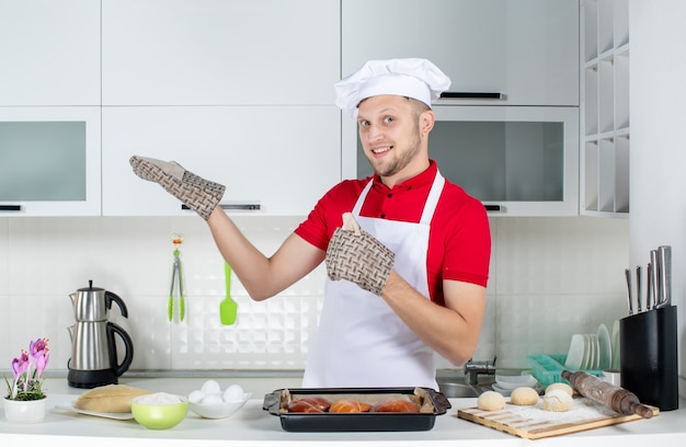 Vista dall'alto dello chef maschio fiducioso che indossa il supporto in piedi dietro il tavolo con le uova di pasticceria grattugia su di esso e mostra qualcosa sul lato destro nella cucina bianca