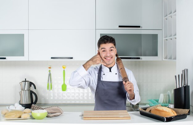 Вид сверху уверенного в себе молодого повара в униформе, держащего терку и делающего жест "позвони мне" на белой кухне
