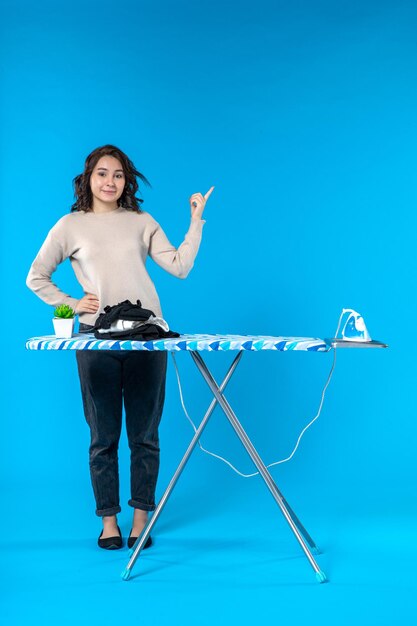 Top view of confident young girl standing behind the ironing board and pointing back on blue wave background