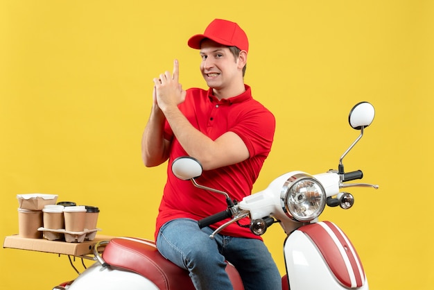 Top view of confident young guy wearing red blouse and hat delivering orders making gun gesture on yellow background