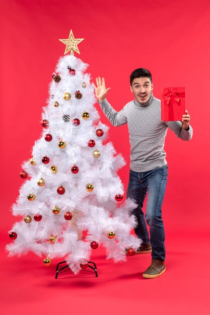 장식 된 흰색 크리스마스 트리 근처에 서서 그의 선물을 들고 빨간색에 다섯을 보여주는 확신 젊은 남자의 상위 뷰