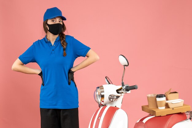 Top view of confused courier girl in medical mask standing next to motorcycle with coffee cake on it on pastel peach color background