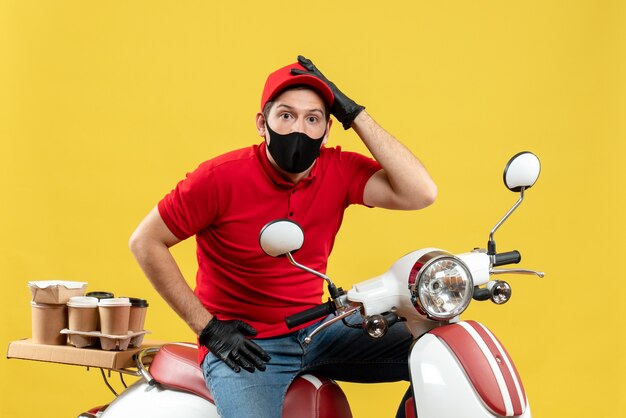 Top view of confused courier guy wearing red blouse and hat gloves in medical mask delivering order sitting on scooter