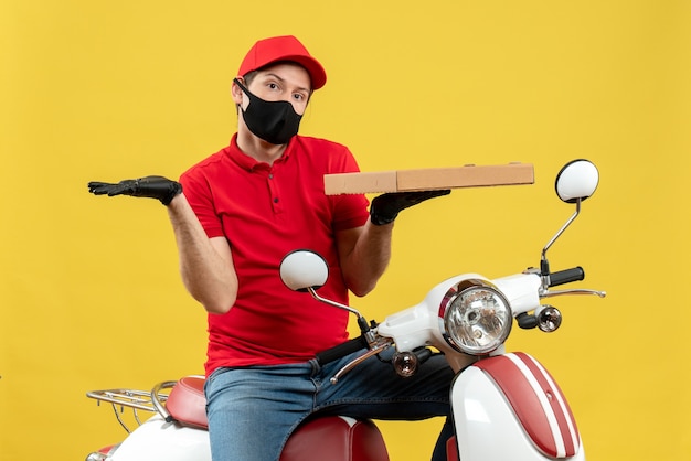 Top view of confused courier man wearing red blouse and hat gloves in medical mask holding order showing something on the left side