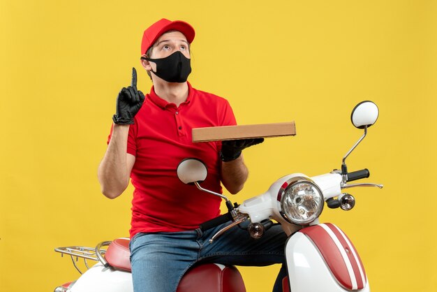 親指を上にして注文を保持しているスクーターに座っている医療マスクで赤いブラウスと帽子の手袋を身に着けている混乱した宅配便の男の上面図