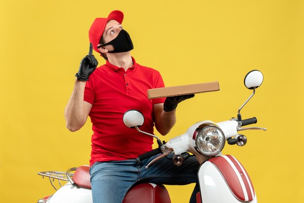 Top view of confused courier man wearing red blouse and hat gloves in medical mask sitting on scooter showing order pointing up