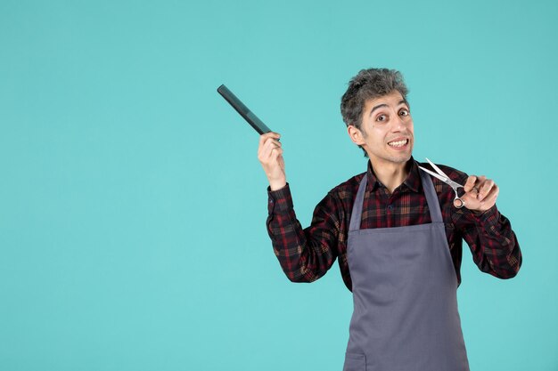 Top view of confused male barber wearing gray apron and holding comb scissor on blue color background