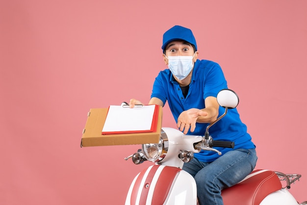 Vista dall'alto del fattorino maschio confuso in maschera con cappello seduto su scooter che consegna ordini in possesso di documento su sfondo pesca peach