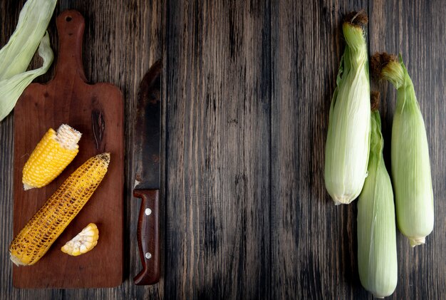 Top view of cooked corns on cutting board with knife and uncooked corns on wood with copy space