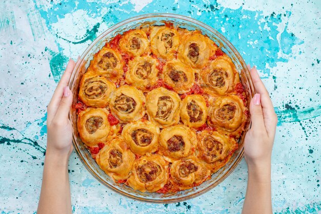 Top view of cooked dough meal with minced meat and tomato sauce inside glass pan taking by female on bright-blue desk, cooking bake food meat dough