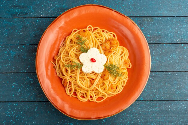 Вид сверху приготовленной итальянской пасты, вкусной еды с зеленью внутри оранжевой тарелки на синей деревянной деревенской поверхности