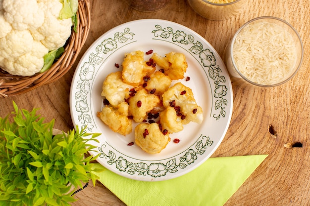 Top view of cooked sliced cauliflower inside plate with greens and raw rice on the cream wooden surface