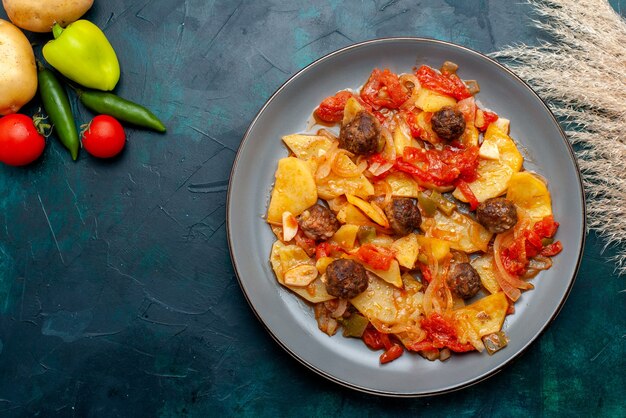 Top view cooked sliced potatoes with meatballs and cooked vegetables on dark-blue desk.