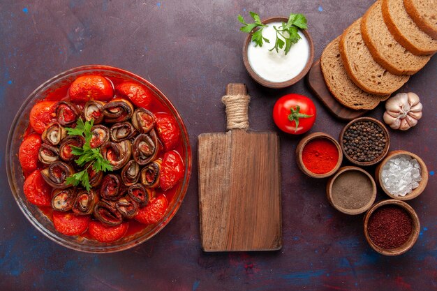 Top view cooked vegetable meal tomatoes and eggplants with seasonings and bread loafs on dark desk