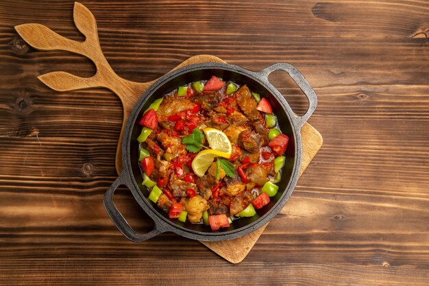 Top view cooked vegetable meal with meat and sliced bell-peppers inside pan on brown wooden surface