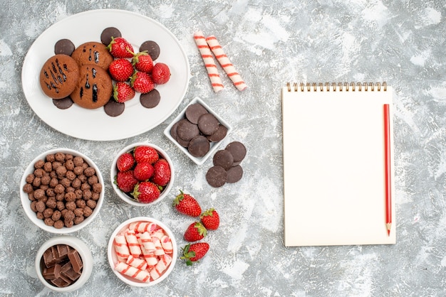 무료 사진 사탕 딸기 초콜릿 시리얼과 회색-흰색 테이블에 연필로 노트북 타원형 접시 그릇에 상위 뷰 쿠키 딸기와 둥근 초콜릿
