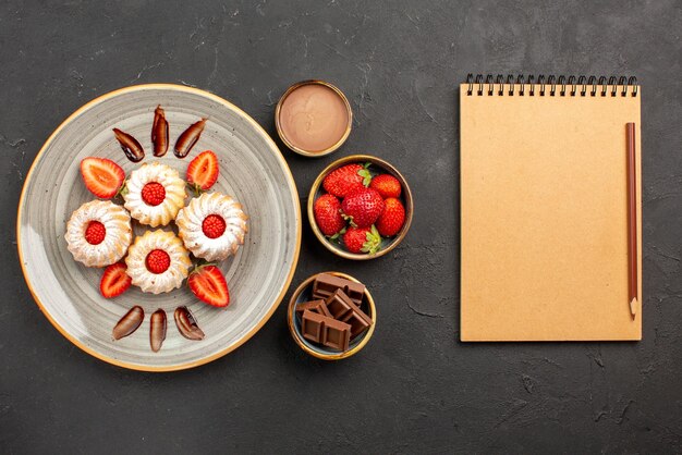 노트북과 연필 옆에 딸기 초콜릿과 초콜릿 크림이 담긴 그릇 옆에 하얀 접시에 딸기가 있는 상위 뷰 쿠키와 딸기 쿠키
