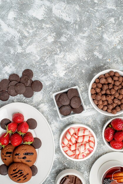 Вид сверху печенье клубника и круглые шоколадные конфеты на овальной тарелке, миски с конфетами клубника, шоколадные хлопья и чашка чая внизу серо-белого стола