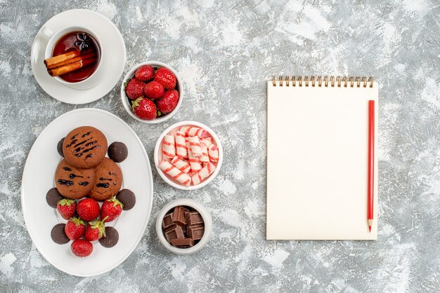 Top view cookies strawberries and round chocolates on the oval plate surrounded with bowls of candies strawberries chocolates cinnamon tea and notebook pencil on the grey-white table