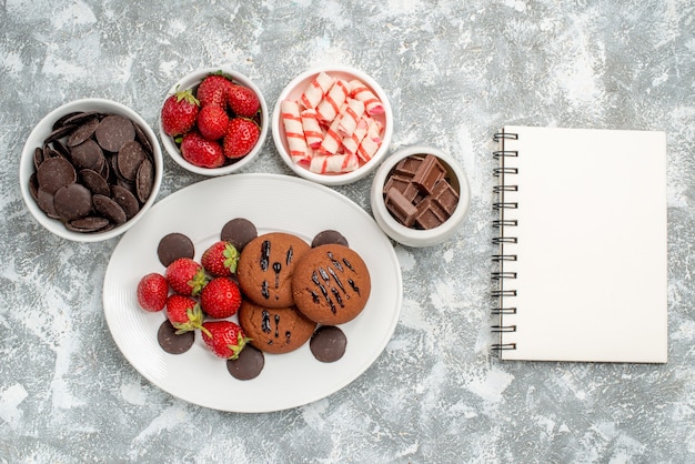 Top view cookies strawberries and round chocolates on the white oval plate surrounded bowls with candies strawberries and chocolates and a notebook on the grey-white table