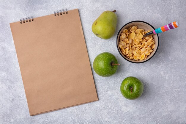 Top view  copy space beige notepad with green apples  cornflakes in a bowl with a spoon and pear on a white background